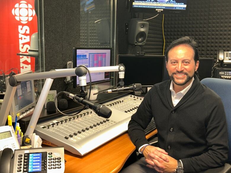 A man sits inside a control room for a radio studio. He is surrounded by monitors, a microphone and a sound board. 