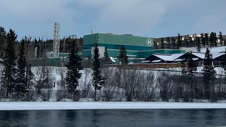 A large green building marked with an 'H' is seen in winter, on the other side of a partially-frozen river.