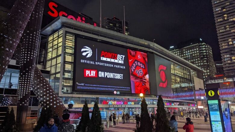 People mill about outside Toronto's Scotiabank Arena at night, with PointsBet Canada branding on a giant digital sign on the arena in the background.