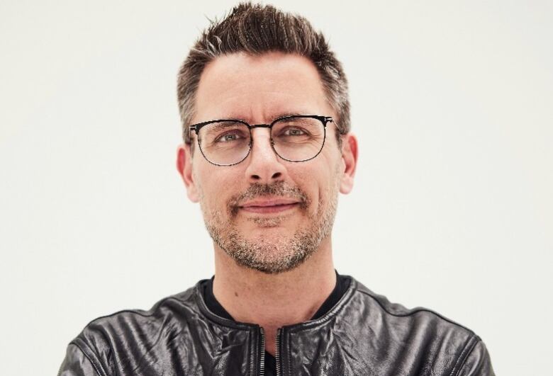 A headshot of a fit, middle-aged Caucasian man with glasses and salt & pepper hair and facial hair, wearing a black leather jacket, with arms crossed.