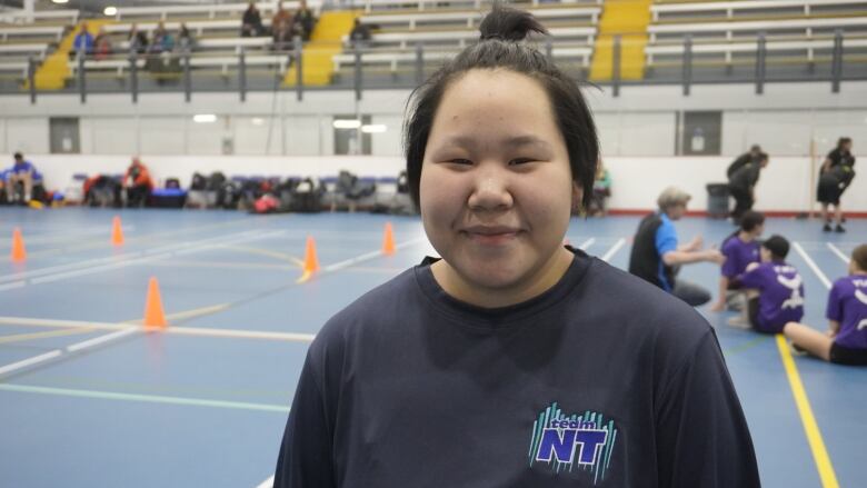 A girl smiles, standing in a gym with a blue floor. 