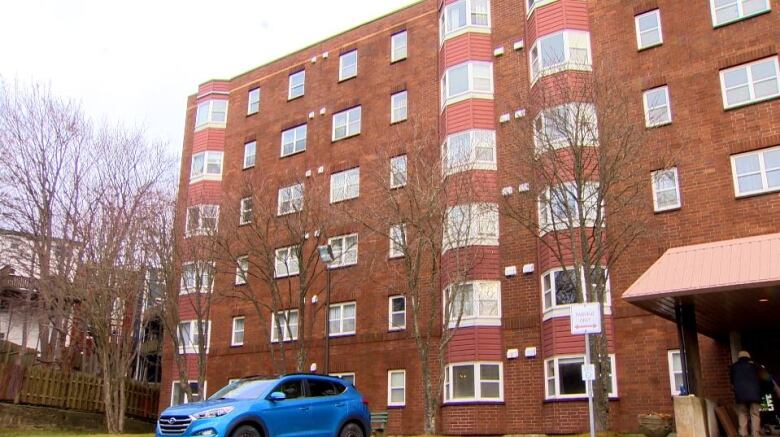 A tall brick building with a blue car parked in front. To the right are two people going through a door. 