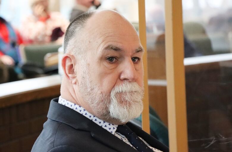 A man with a white beard and moustache wearing a dark blue suit looks toward the camera.