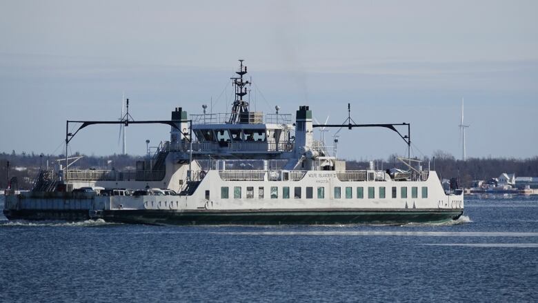 A large white and green ferry with a flat deck is shown sailing toward an island where homes and wind turbines can be seen.