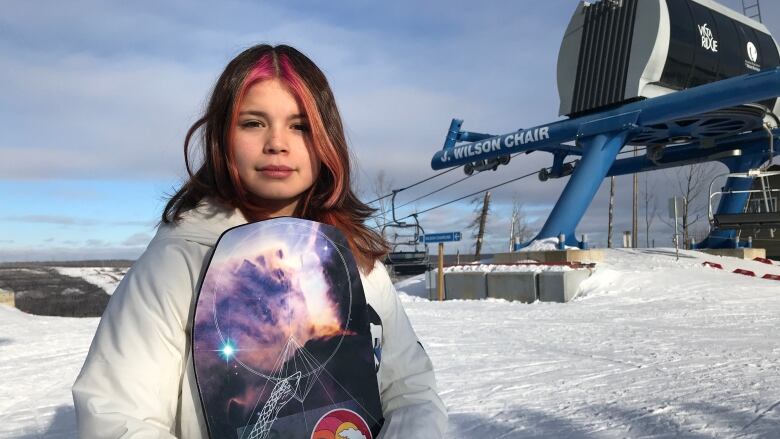 A girl stands holding a snowboard at the top of a ski hill. There's a chairlift in the background.