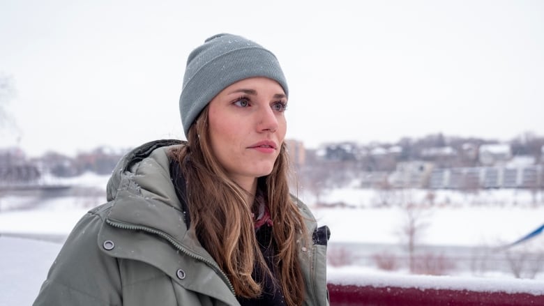 A woman wearing a tuque and parka stands outside during wintertime and gazes beyond the camera. 