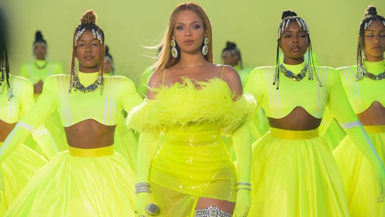 Beyonce, centred, woman in yellow dress stands fiercely and confidently surrounded by several other women in similar florescent yellow tops and skirts and crystal accessories