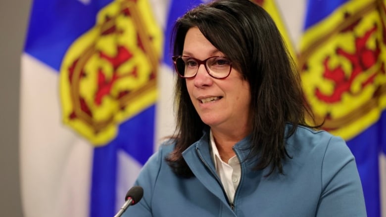 A woman with dark hair and glasses sits at a podium.