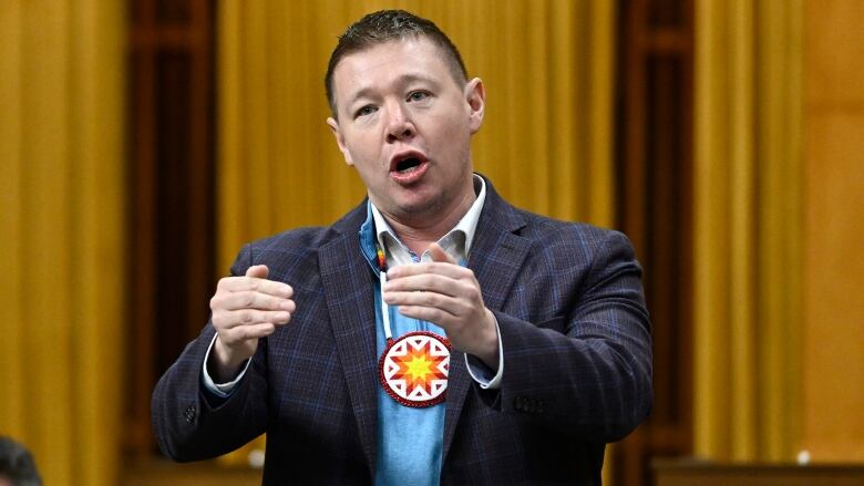 A man in a blue suit with an Indigenous symbol on a necklace gestures with his hands as he speaks.