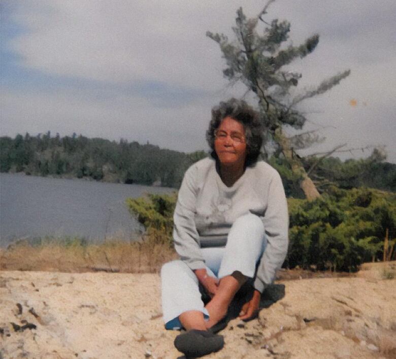 A woman with black hair and glasses, wearing a grey sweatshirt and blue jeans, sits on a rocky shoreline with a lake and evergreen trees behind her.