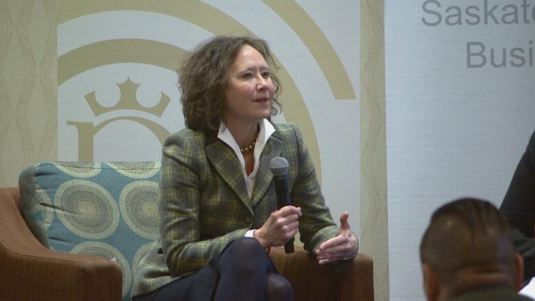 A woman in a suit sits in an armchair holding a microphone in one hand while speaking at a public event.