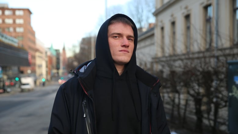 A man wearing a black hoodie poses for a photo on an empty street, with a line of trees and buildings behind him.