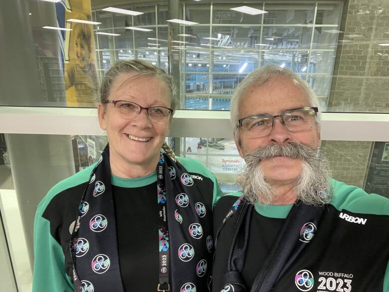 A man and woman pose for a photo. They are wearing green and black volunteer sweaters for the Arctic Winter Games. 