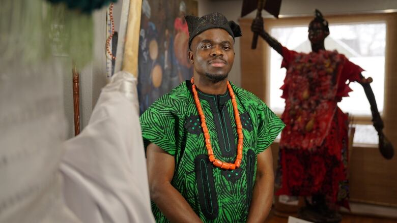 African man stands between two statues. One is wearing a red tunic and holding a double bladed battle axe, the other is dressed in white. The man is wearing a dark green dashiki, an coral bead necklace and a black cap.