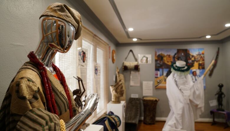 A room filled with artifacts and artwork.  Two tall statues are opposite each other. One wears a long white robe and white hat, that has green trim on it. It holds a long wooden stick. The other wears a tan Dashiki and matching cap. 