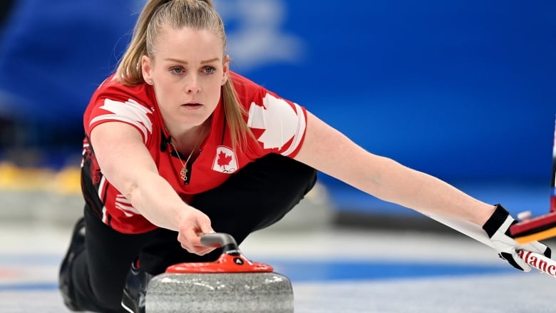 A female curler pushes a stone forward with her right hand as she slides on the ice on her left foot with her left knee bent forward and her right knee touching the ice.