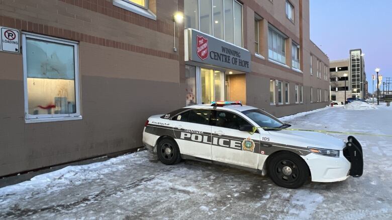 A police car with yellow caution tape tied to its side mirror, blocks the snowy parking lot of a building. A name above the door to the building says Winnipeg Centre of Hope.