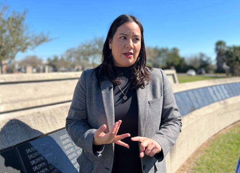 A woman with long dark hair and wearing a blazer gestures while standing in a park.