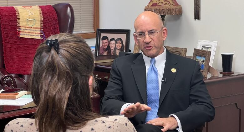 A bald man wearing glasses and a suit sits in a chair in an office talking to a woman.