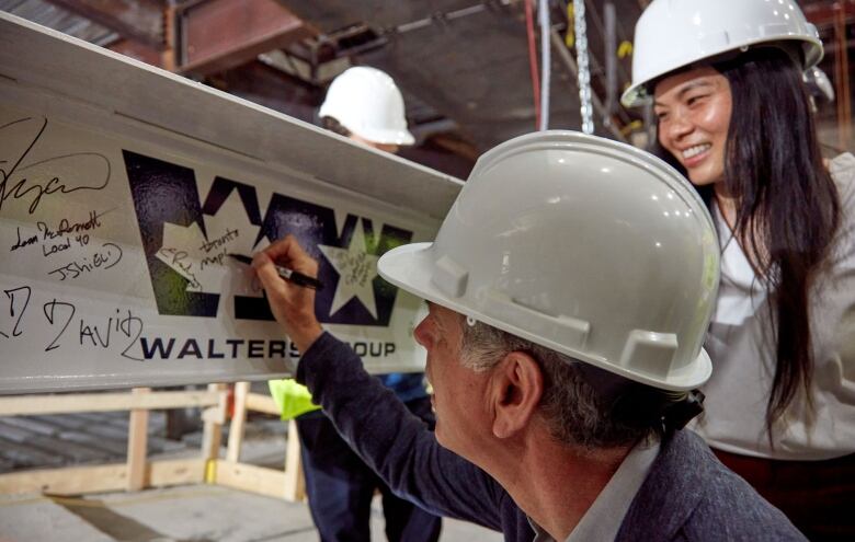 Man wearing a hard hat signs a beam while a woman looks on.