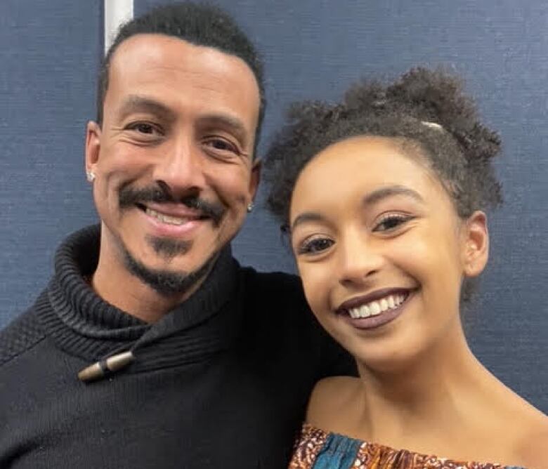 A young Black man with a moustache and goatee stands smiling, wearing a black sweater, next to a young Black woman with dark curly hair who is wearing a colourful printed off the shoulder shirt in shades of teal, yellow and maroon.