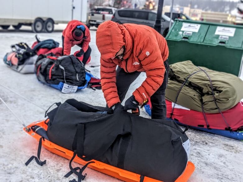 A man, wearing a winter coat with the hood on, is bent over a long black bag, on top of a sled. He is surrounded by other sleds and one other participant. 