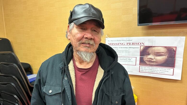 A man with a leather cap, red t-shirt and black winter jacket is pictured in a hotel room in front of a missing persons poster.