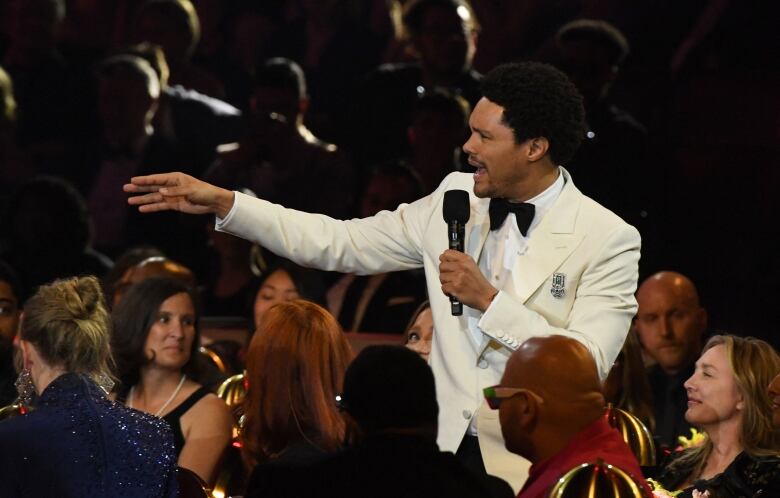 A man in a white tuxedo smiles broadly and gestures towards the left. He is standing amongst well dressed people seated in a banquet hall.