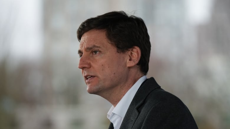 An earnest-looking young man in a grey suit and white shirt speaks in front of a blurred-out background.