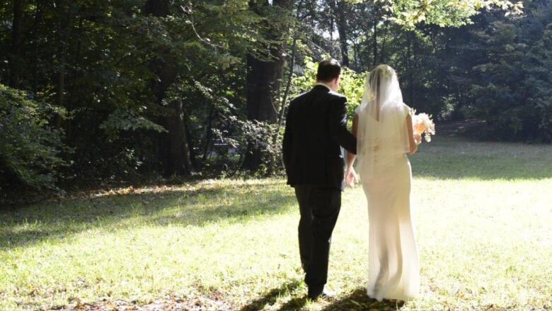 A woman and man walk away from the camera on grass, dressed in wedding attire.