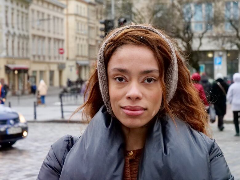 Woman with reddish brown hair and wearing a knit headband and a blue winter jacket stands on a city street.