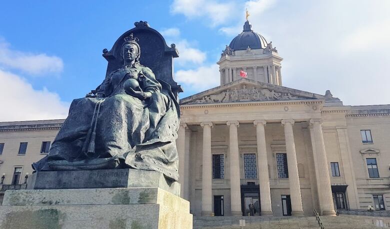 A statue of a woman seated on a throne stands in front of the Manitoba legislative building, which has stairs leading to several stone columns and a dome topped by a golden statue.