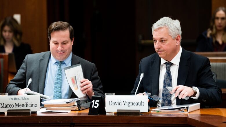 Two men in suits, with binders of papers in front of them, sit at a table.