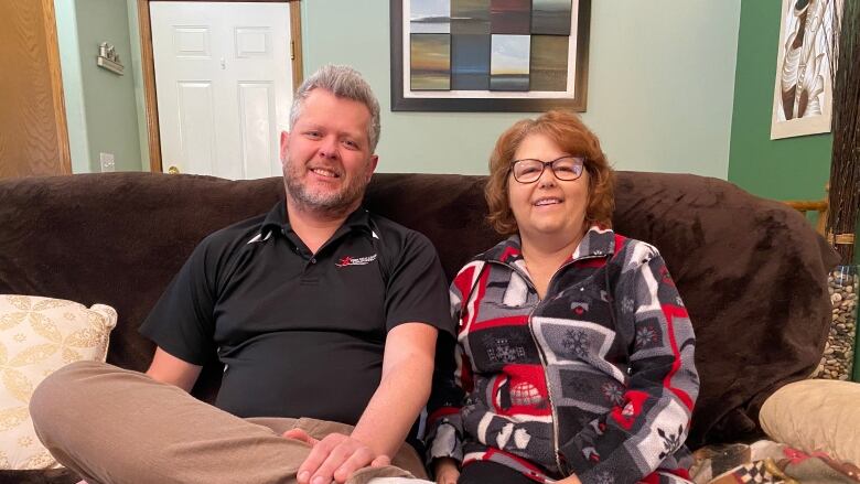 A man in a black t-shirt and brown trousers sits next to a woman on a couch.