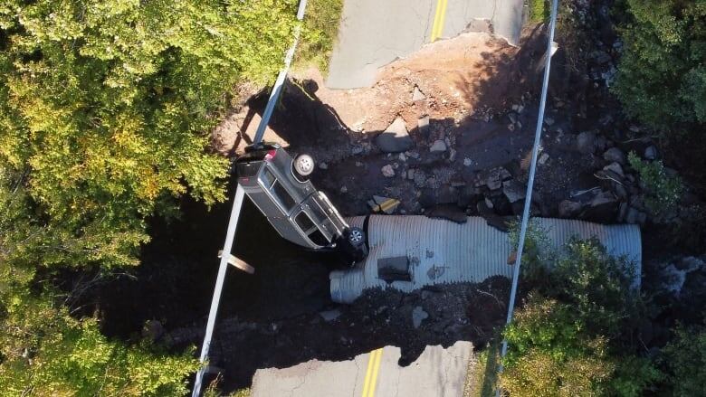 A dark-coloured SUV rests precariously on a culvert and guardrail along a damaged road.