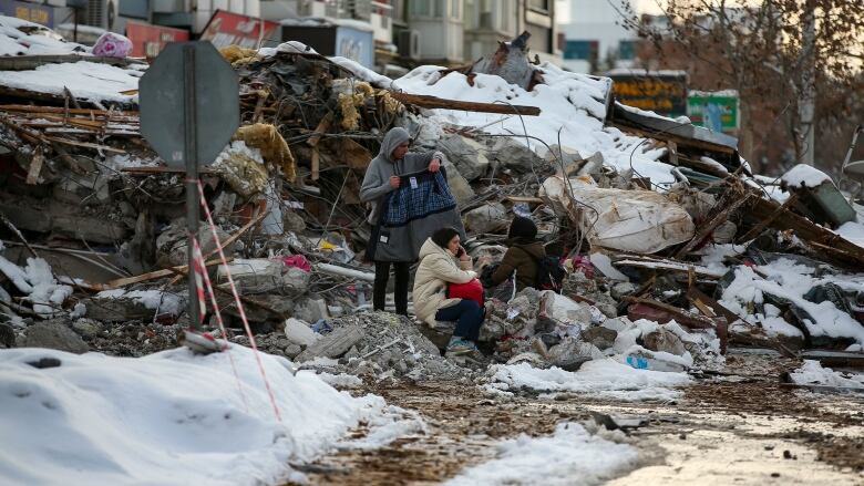 A man stands on a pile of snow-covered debris, holding a jacket, while a woman is sitting in front of him and holding a child.