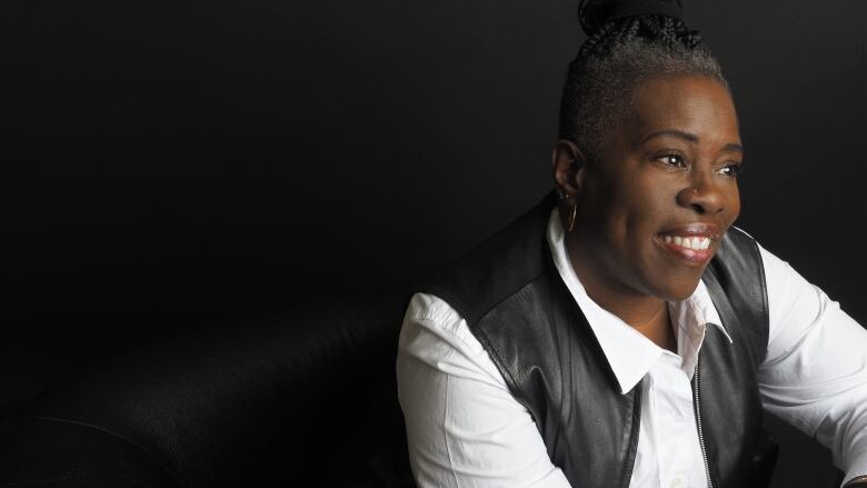 A Black woman with swept-up, grey braided hair and wearing a nose piercing, a white open-collared shirt and a black leather vest looks off-camera.