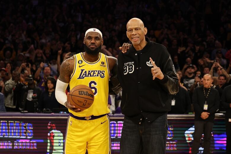 A basketball player wearing a yellow Los Angeles Lakers uniform holds a ball as he poses to a picture with taller man wearing black clothes. 