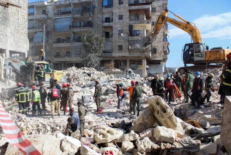 Several workers and onlookers are shown standing in rubble in front of a multi-storey building as a crane operates nearby.