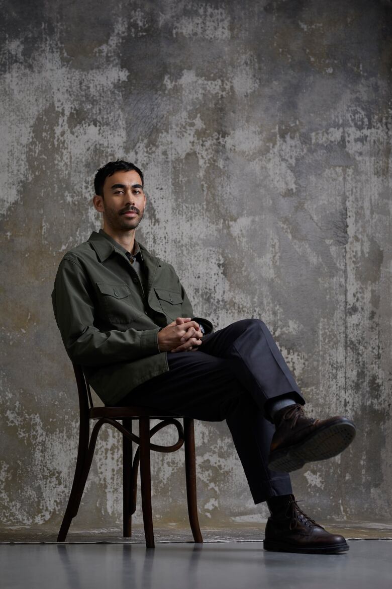 Antoine Bourges sitting in a chair, legs crossed and arms folded, against a concrete-style backdrop.