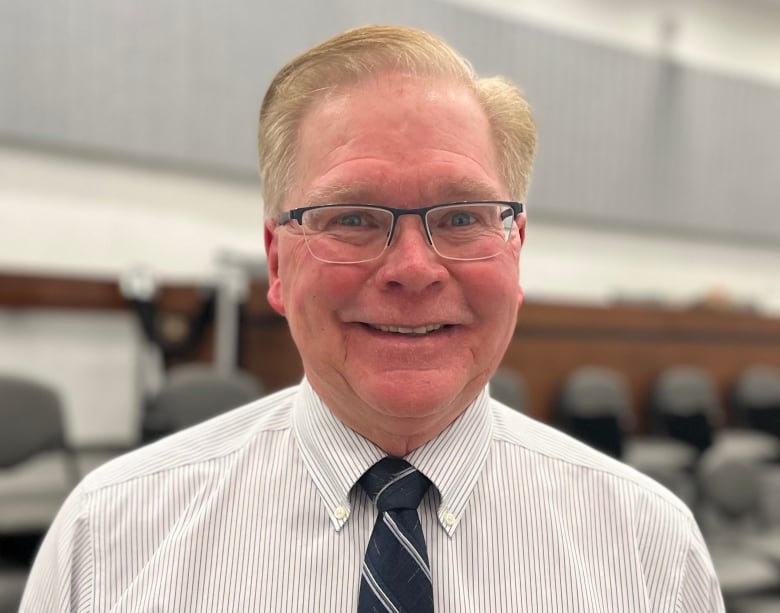 A portrait of a middle-aged man in a striped shirt, navy and white striped tie and glasses. 