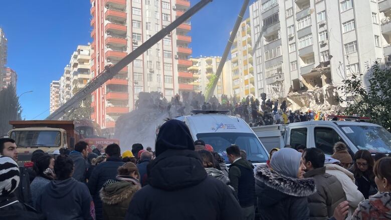 A large group of onlookers gather behind construction vehicles, as others dig through a large pile rubble following a building collapse. In the background, some apartment buildings still stand.