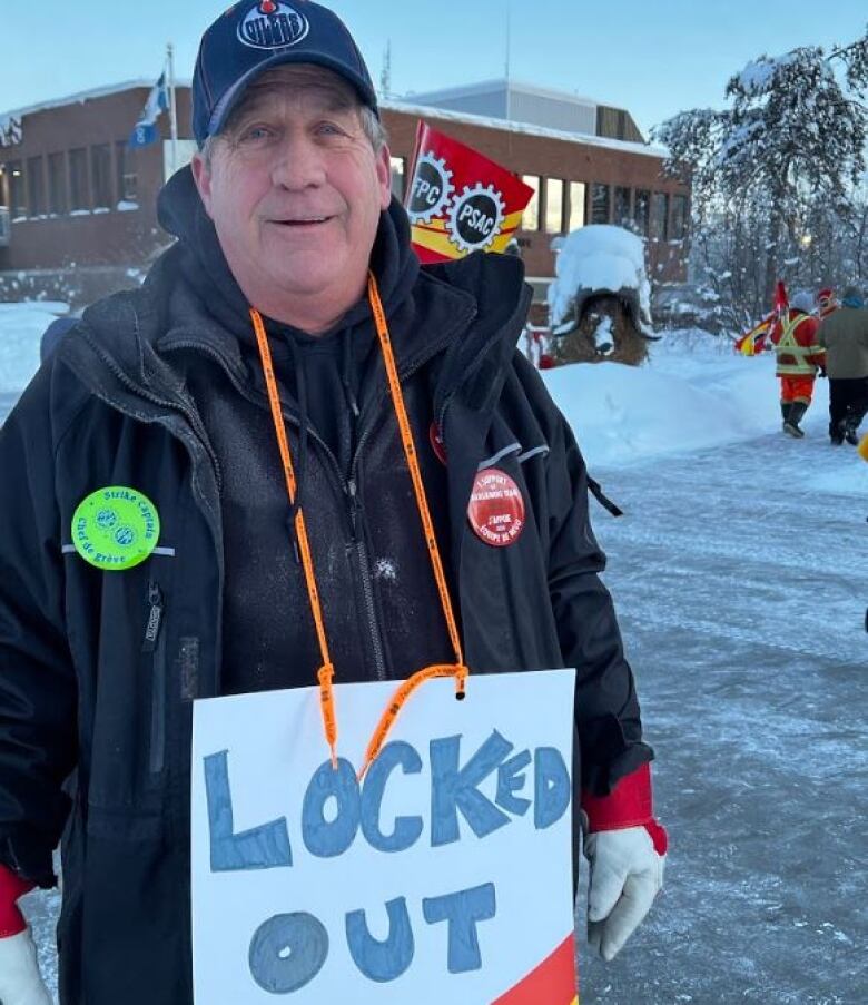 A portrait of a smiling, red-cheeked man outside in the snow with a sign around his neck that says 