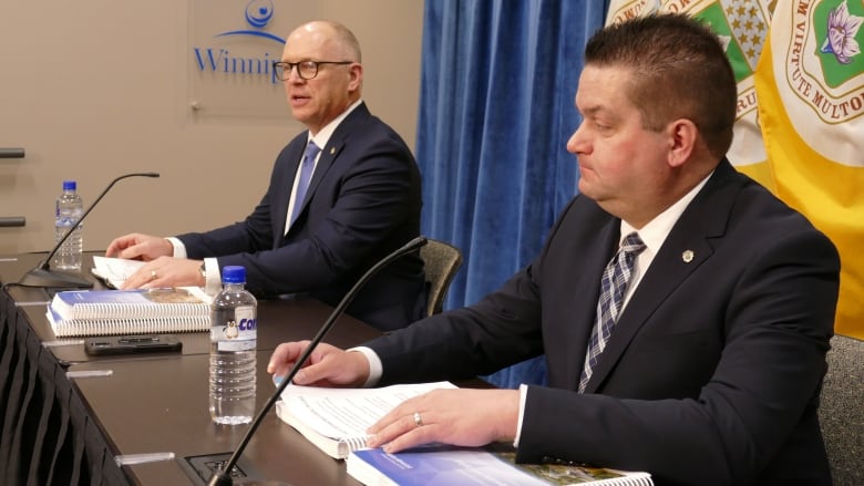 Two men in suits at a table in front of flags.