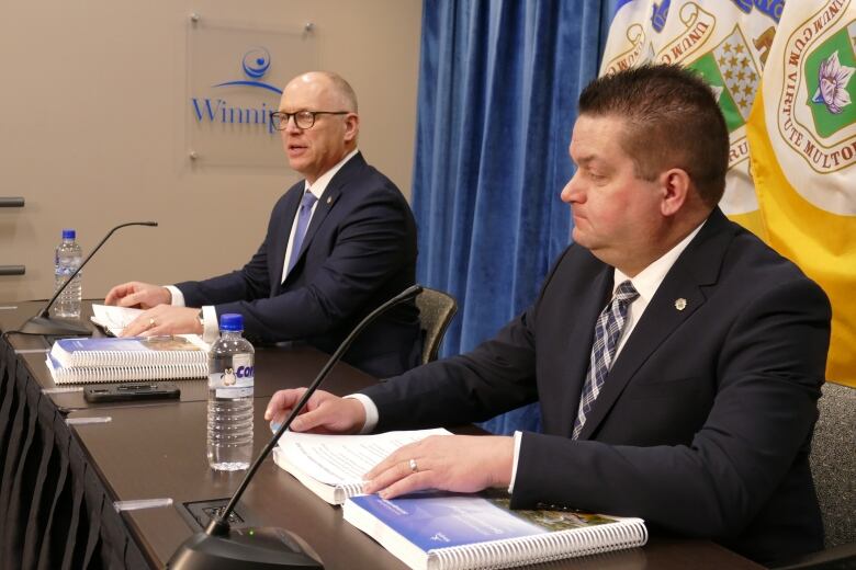 Two men in suits at a table in front of flags.