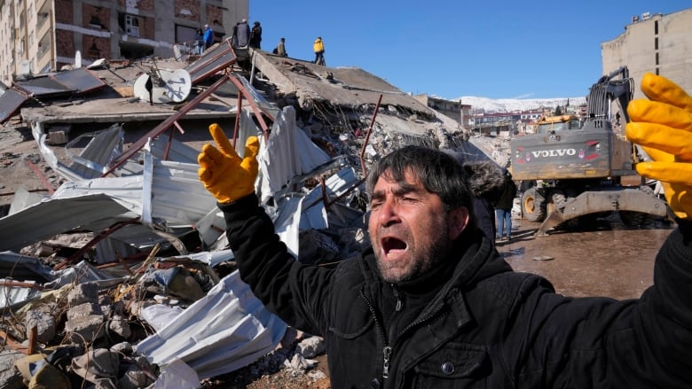 A man close to the camera cries out and raises his yellow-gloved hands, in front of a collapsed building, which people are standing on top of in the background.