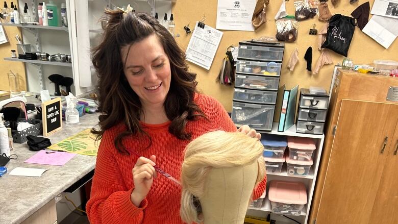 A woman with brown hair and a bright red sweater works on a wig while holding a can of hair spray.