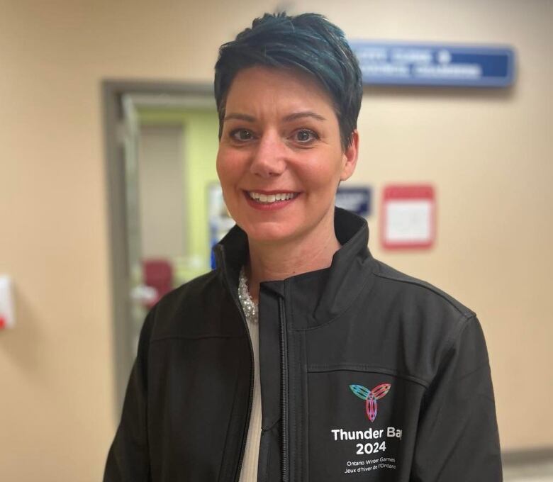 A woman with short blue and black hair stands in a hallway, smiling.