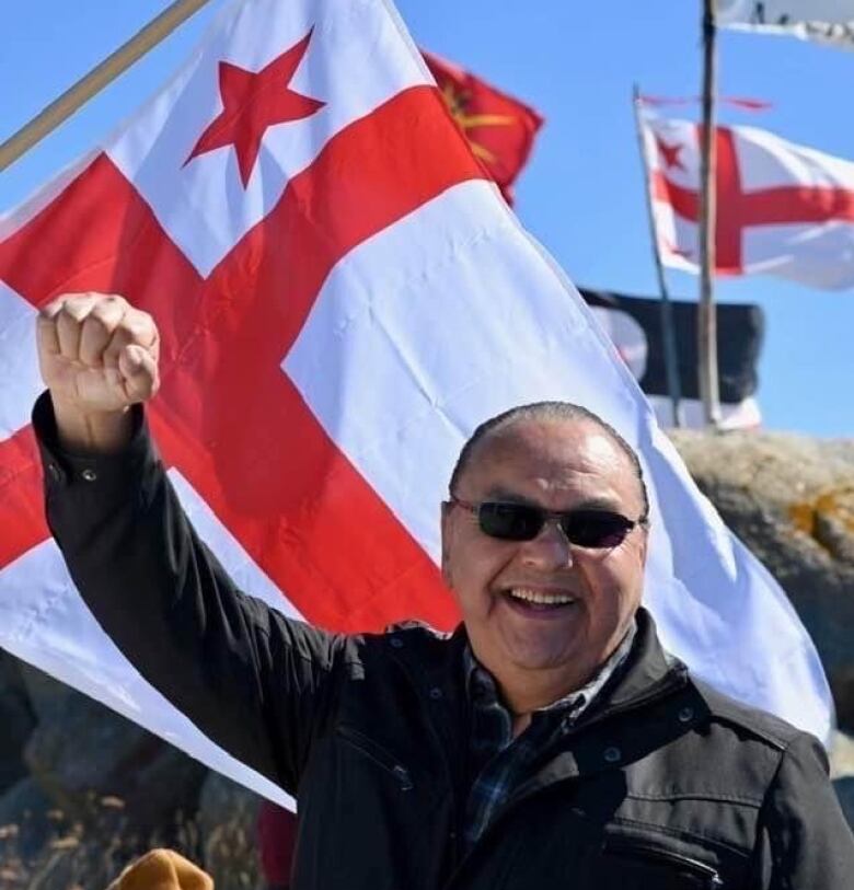 A man raises a fist on a sunny day. In the background is a Mi'kmaq flag.