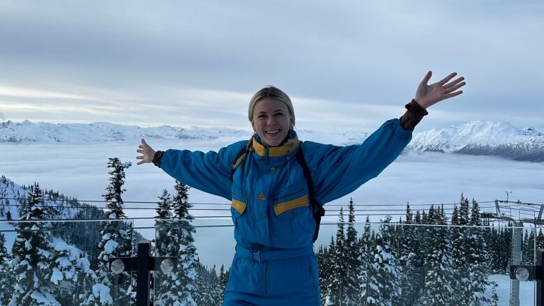 A woman in a blue and yellow snow suit stands with arms outstretched against a snowy mountain backdrop. 